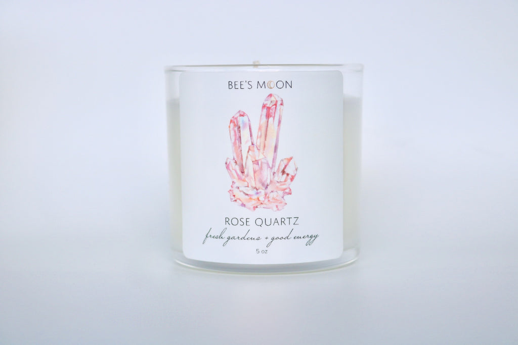 A cluster of strawberry crystal quartz painted on a label of the rose quartz candle on an empty table with bright white space around it. The candle is unlit.